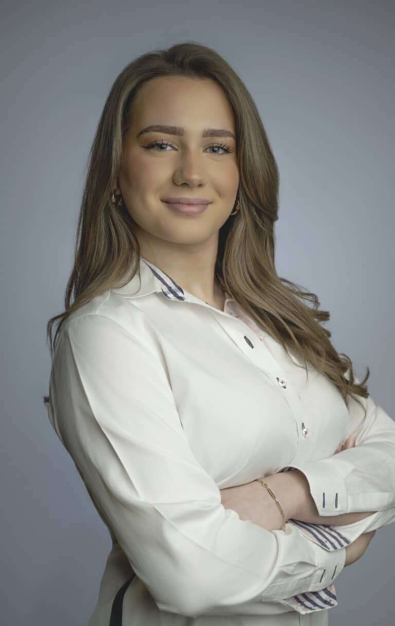 Portrait of a smiling woman with long light brown hair wearing a white shirt with striped cuffs, arms crossed against a gray studio background.