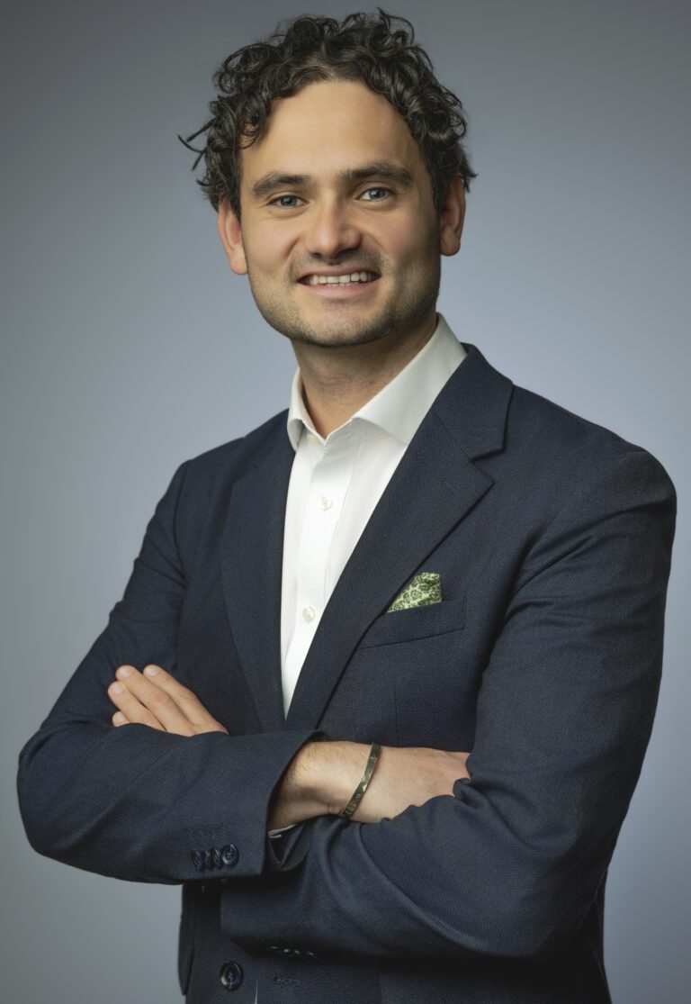 Smiling man in a navy blazer with white shirt, arms crossed, posing against a gray backdrop.