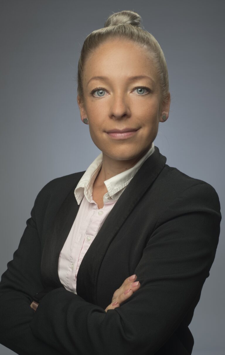 Professional head-and-shoulders portrait of a confident woman with blonde hair in a black blazer and white shirt, arms crossed against a gray gradient background.